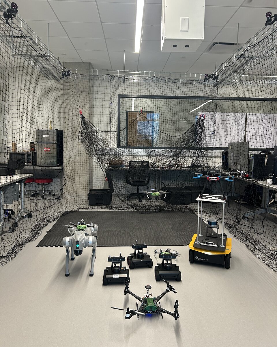 The testing arena, enclosed by safety netting with motion-capture cameras along the top, with a quadruped, three ground robots, a Jackal robot, and two drones on the floor.